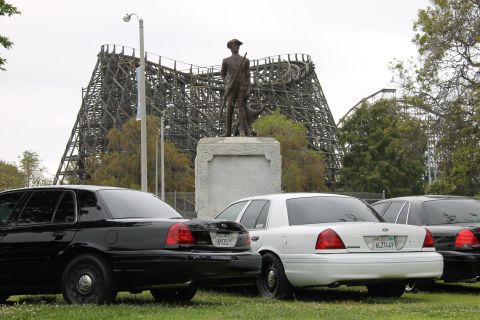2004 Ford Crown Victoria - A trio of police sedans are parked on a grassy area, with a large wooden roller coaster and a statue visible behind them.