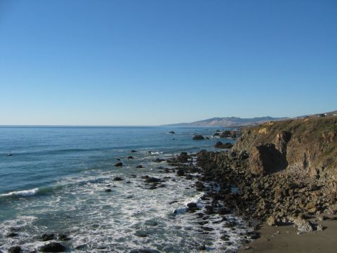 2002 Ford Taurus - Rocky Pacific coastline under a clear blue sky, with surf breaking over dark boulders, a sandy beach at right and headlands receding into t…