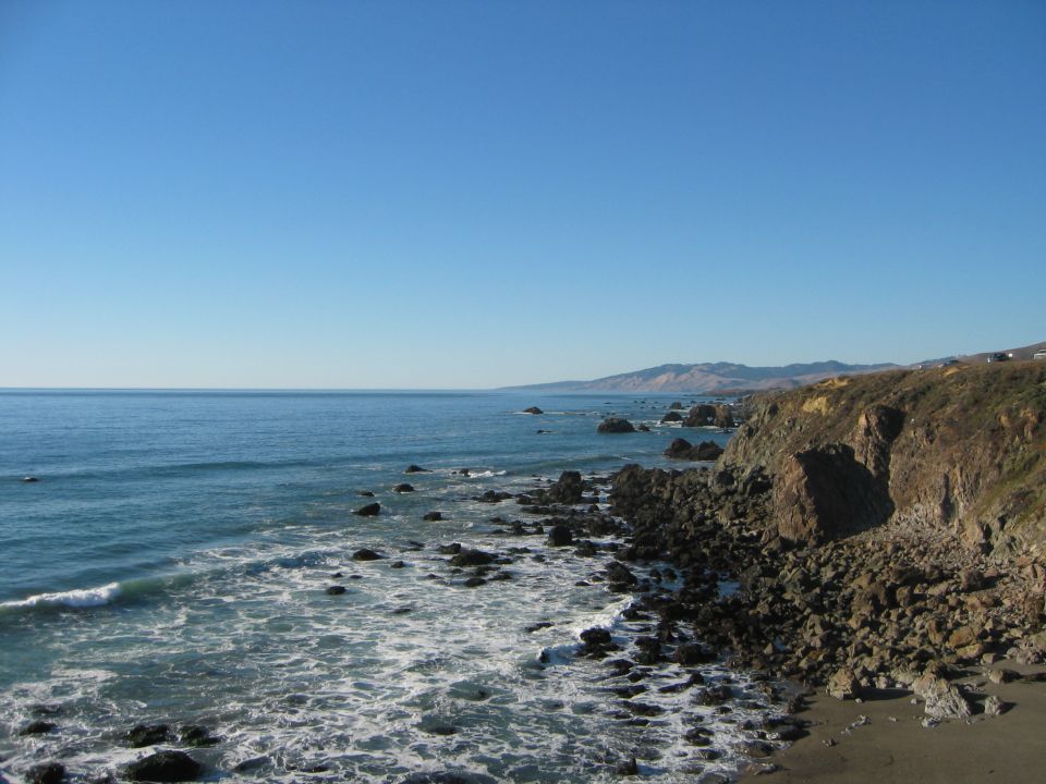 Rocky Pacific coastline with surf, dark boulders and distant headlands under blue sky