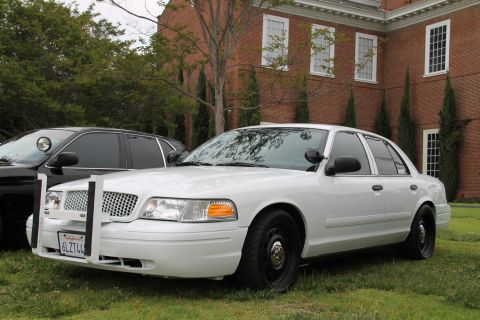 2004 Ford Crown Victoria - A white 2004 Ford Crown Victoria police car is parked on grass in front of a brick building, with another police car visible to its left.