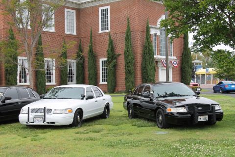 2004 Ford Crown Victoria - Two Ford Crown Victoria police cruisers are parked on the lawn outside a brick building.