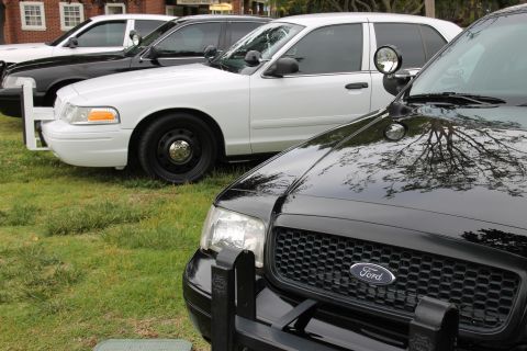 2004 Ford Crown Victoria - Several police cars, including a black Ford and a white Ford, are parked in a row on a grassy area.