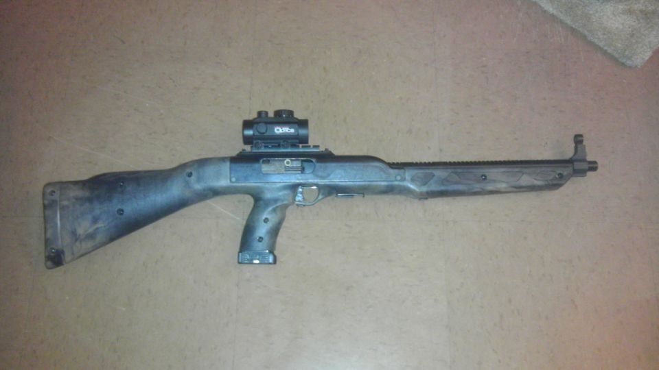 Black Hi-Point 995 Carbine with red dot sight, lying on a textured floor