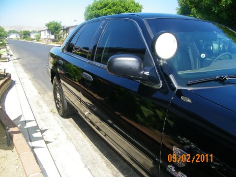 2003 Ford Crown Victoria - A black 2003 Ford Crown Victoria sedan is seen from the side, parked on a street with a spotlight attached.
