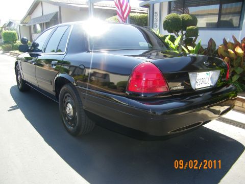 2003 Ford Crown Victoria - Rear quarter of a black Ford sedan on steel wheels parked near a flag and shrubs.
