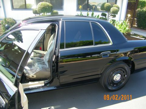 2003 Ford Crown Victoria - Rear driver door open on a black sedan showing a tan rear seat partly covered in plastic.