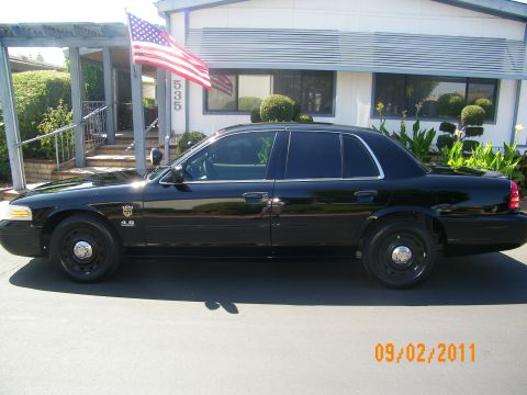 2003 Ford Crown Victoria - Side view of a black Crown Victoria P71 parked by a home with US flag; 4.6 badge and blacked-out steel wheels visible.