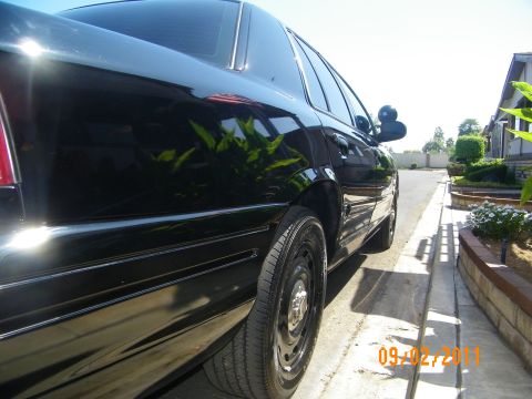 2003 Ford Crown Victoria - Low-angle view down the driver side of a black Crown Victoria along a curb, black steel wheels in frame.