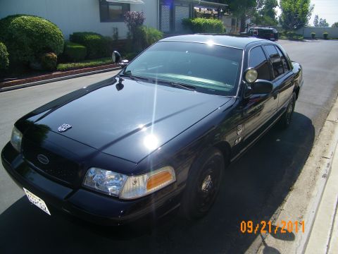 2003 Ford Crown Victoria - Dark Crown Victoria photographed front quarter in a driveway, Ford oval grille badge and pillar spotlight visible.