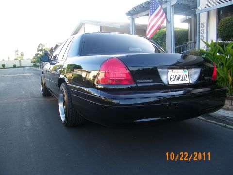 2003 Ford Crown Victoria - Tail end of a black Crown Victoria Police Interceptor, plate 6SQR002, by a flagged house.