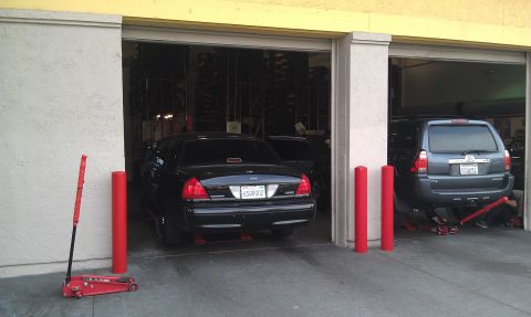 2003 Ford Crown Victoria - Two vehicles in open garage bays beside red posts and a hydraulic floor jack