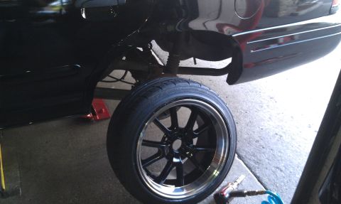 2003 Ford Crown Victoria - Multi-spoke black wheel on a performance tire propped next to a lifted car