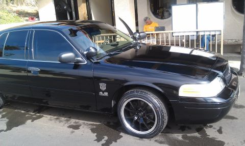 2003 Ford Crown Victoria - Glossy black Crown Victoria parked under a carport, hood ajar, 4.6 fender badge and black wheels visible