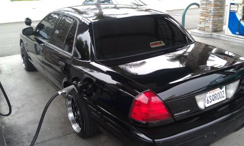 2003 Ford Crown Victoria - Black Crown Victoria at a fuel pump with nozzle inserted, red taillights and dark tint
