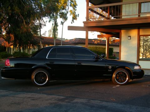 2003 Ford Crown Victoria - Driver profile of a tinted black Crown Victoria, autumn trees and stucco overhang behind.