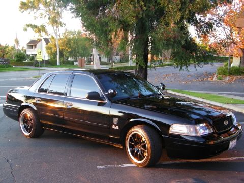 2003 Ford Crown Victoria - Glossy black Crown Victoria parked at curb, autumn leaves scattered on the pavement.