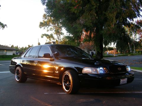 2003 Ford Crown Victoria - Front three-quarter of a black Crown Victoria with lit headlights in suburban parking lot.