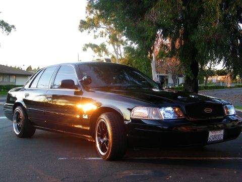 2003 Ford Crown Victoria - Black Crown Victoria angled in a parking lot at sunset, headlights glowing, fender emblem visible.