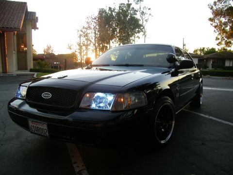 2003 Ford Crown Victoria - Black Crown Victoria with HID-style headlights glowing, sun setting behind the buildings.
