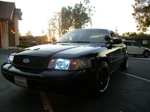 2003 Ford Crown Victoria - Black Crown Victoria at dusk with lit headlights, a silver Mini Cooper parked at right.