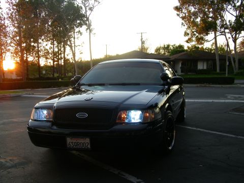 2003 Ford Crown Victoria - Low front-quarter angle of a black full-size Ford sedan, sun flaring through trees behind it.