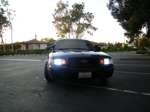 2003 Ford Crown Victoria - Straight-on front shot of a black Ford sedan with headlights lit in an empty lot.