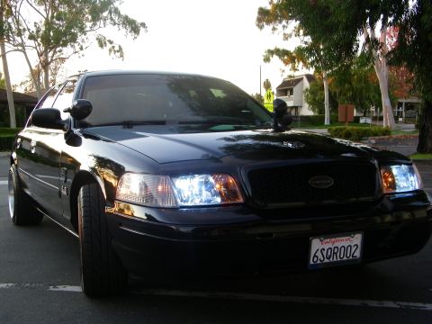 2003 Ford Crown Victoria - Front-quarter angle of a black Ford sedan, headlights on, parked near landscaping.
