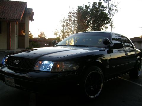 2003 Ford Crown Victoria - Driver-side front quarter of a black Ford sedan parked beside a stucco home at dusk.