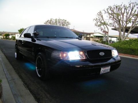 2003 Ford Crown Victoria - Front quarter view of a blacked-out Ford sedan with bright headlights, black wheels, suburban street behind.