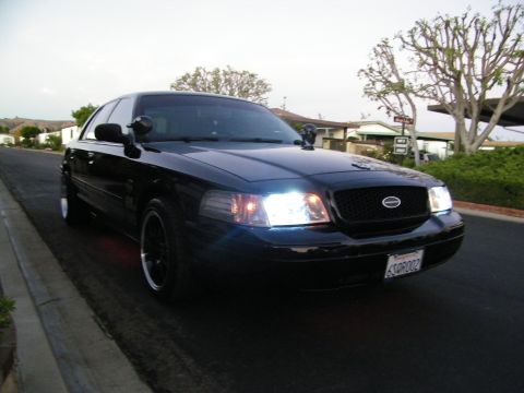 2003 Ford Crown Victoria - Blacked-out full-size Ford sedan shot head-on, HID-style headlights on, parked on a neighborhood road.
