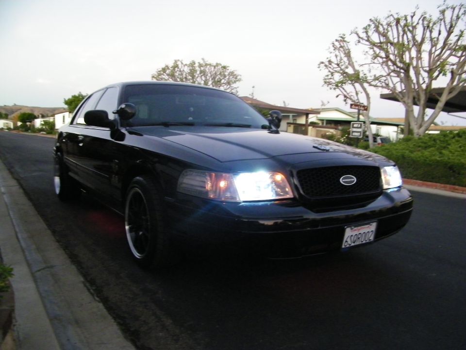 Black Ford sedan front angle with lit headlights, black rims, plate 6SQR002, residential street