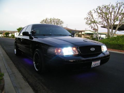 2003 Ford Crown Victoria - Black Ford sedan with purple underglow at the front wheel, lit headlights, suburban backdrop.