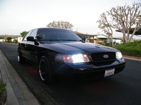 2003 Ford Crown Victoria - Blacked-out Ford full-size sedan at front angle with headlights illuminated on a residential road.
