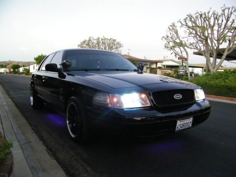 2003 Ford Crown Victoria - Black Ford sedan with purple underglow near the wheel and bright headlights, parked curbside.
