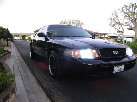 2003 Ford Crown Victoria - Front angle of a black full-size Ford with Ford grille emblem and headlights on, hillside neighborhood.