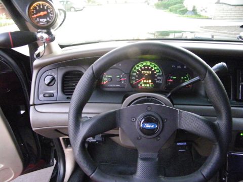 2003 Ford Crown Victoria - View across a Ford dashboard showing a small aftermarket wheel, green gauge cluster, and pillar-mount tach.