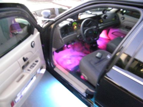 2003 Ford Crown Victoria - Driver door open on a black Ford sedan, pink interior LEDs and tan trim with blue underglow at the sill.