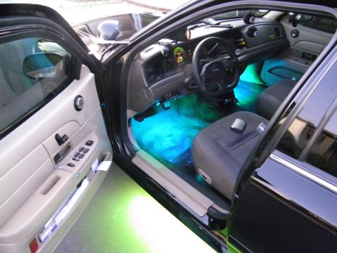 2003 Ford Crown Victoria - Black Ford sedan with door open showing teal interior lighting and green underglow on the pavement.