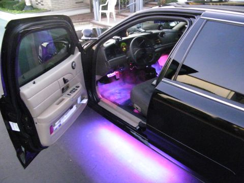 2003 Ford Crown Victoria - Driver door open on a black Ford with purple cabin LEDs glowing onto the ground and tan door trim.