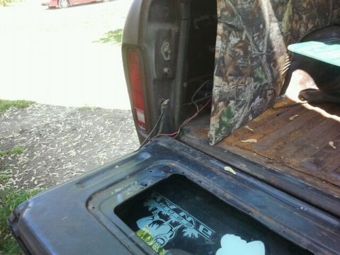 1979 Ford Bronco - View of a truck's open tailgate and bed, a camouflage panel and a Supermotors.net logo, with another vehicle in the distance.