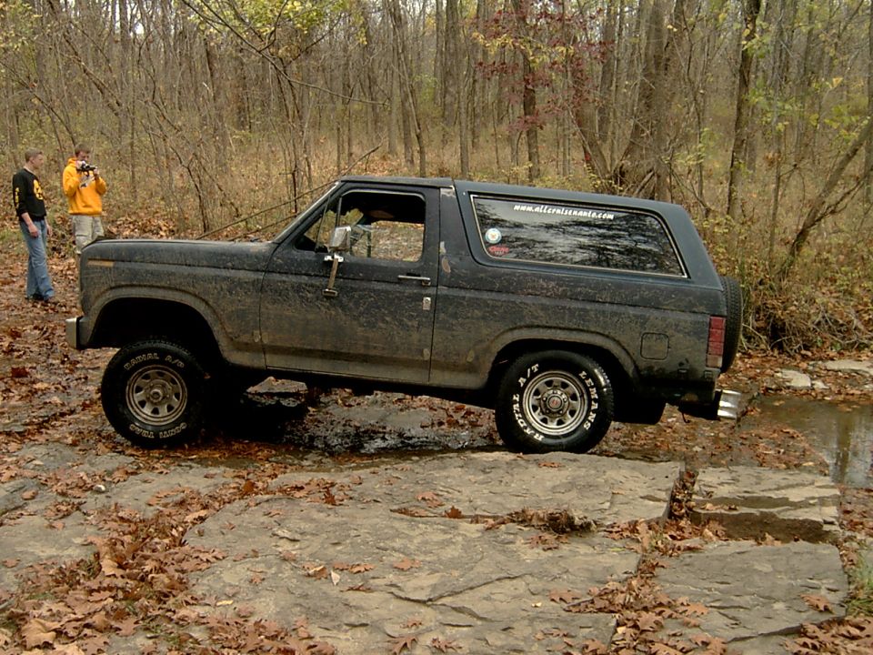Muddy dark Ford Bronco on a rocky wooded trail, driver-side view with hardtop and A/T tires