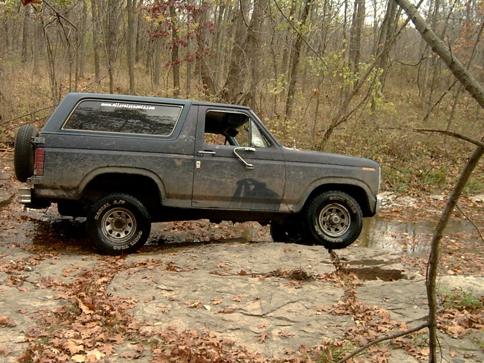 Muddy dark blue Ford Bronco passenger-side profile on rock shelf beside woodland creek