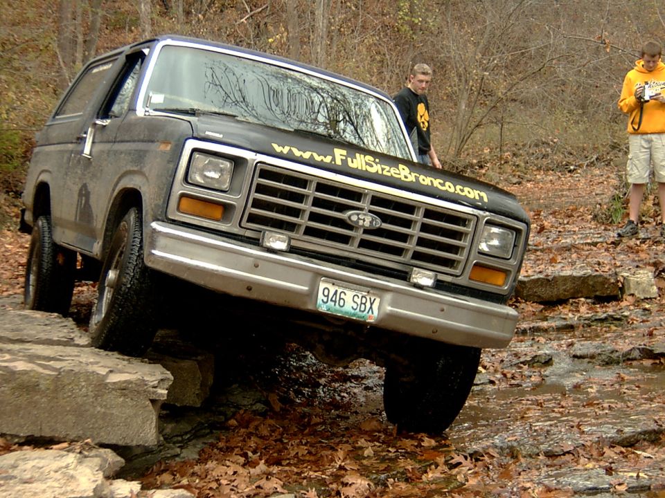 Muddy blue Ford Bronco flexing over rock ledge in creek, FullSizeBronco.com hood decal