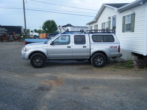 2003 Nissan Frontier - A silver 2003 Nissan Frontier pickup truck with a cap and roof rack is parked on a gravel lot next to a white house.