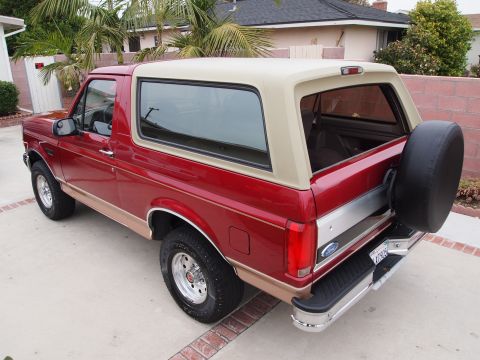 1994 Ford Bronco - This red Ford Bronco with a tan removable top is parked in a driveway with palm trees and a house in the background.