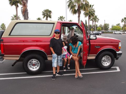 1994 Ford Bronco - A red 1994 Ford Bronco with a tan top is parked in a lot, with its driver's door open and people posing near it.