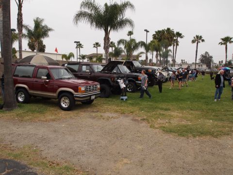 1994 Ford Bronco - A line of trucks and SUVs are parked on a grassy field under a cloudy sky.