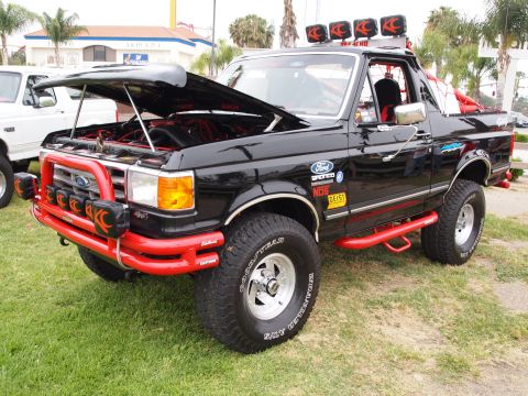 1994 Ford Bronco - This modified black Ford Bronco features a roll cage and KC lights, with a white Ford truck visible beside it.