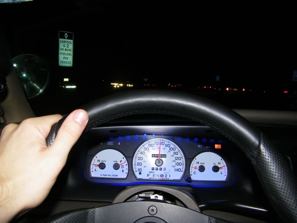 Driver hand on steering wheel at night with blue gauges and carpool lane sign ahead