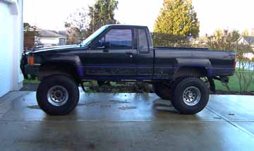 1980 Ford Bronco - This lifted pickup truck is parked outside a building with trees in the background. The truck has large off-road tires.